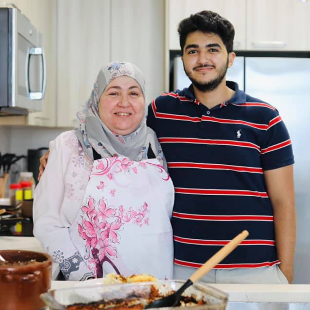 Middle aged woman in a headscarf standing next to her tall, adult son in a home kitchen.