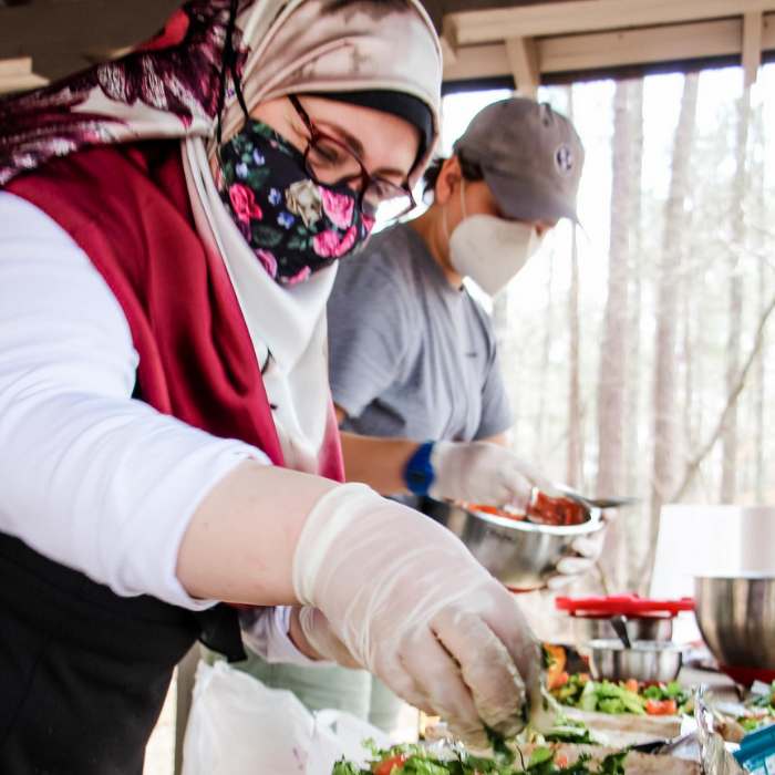 Middle aged woman in glasses, headscarf, and mask with young adult man preparing food outdoors.