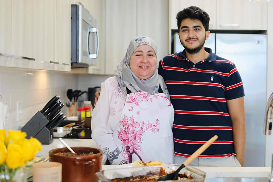 An older woman in a headscarf standing next to her tall, adult son in a home kitchen.