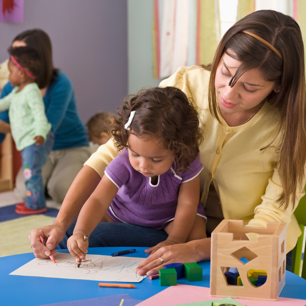 A young, female child care employee sitting at a table with a girl toddler coloring in a child care center classroom. In the background is a second employee playing with two toddlers.
