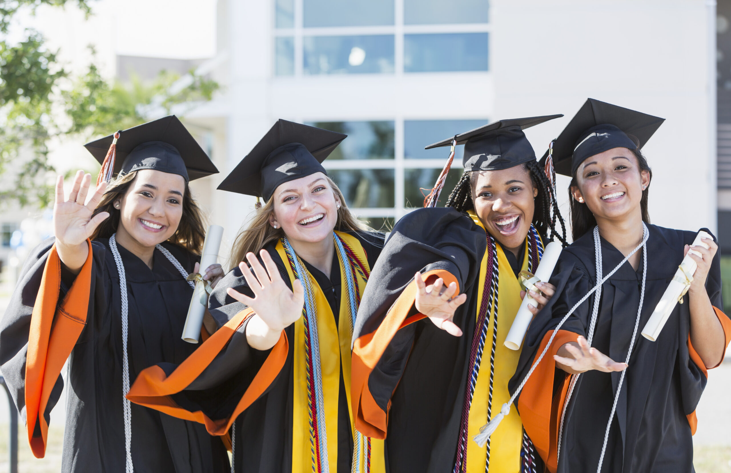 A group of four multi-ethnic young women, 18 years old, wearing graduation caps and gowns, holding diplomas, smiling at the camera, waving.