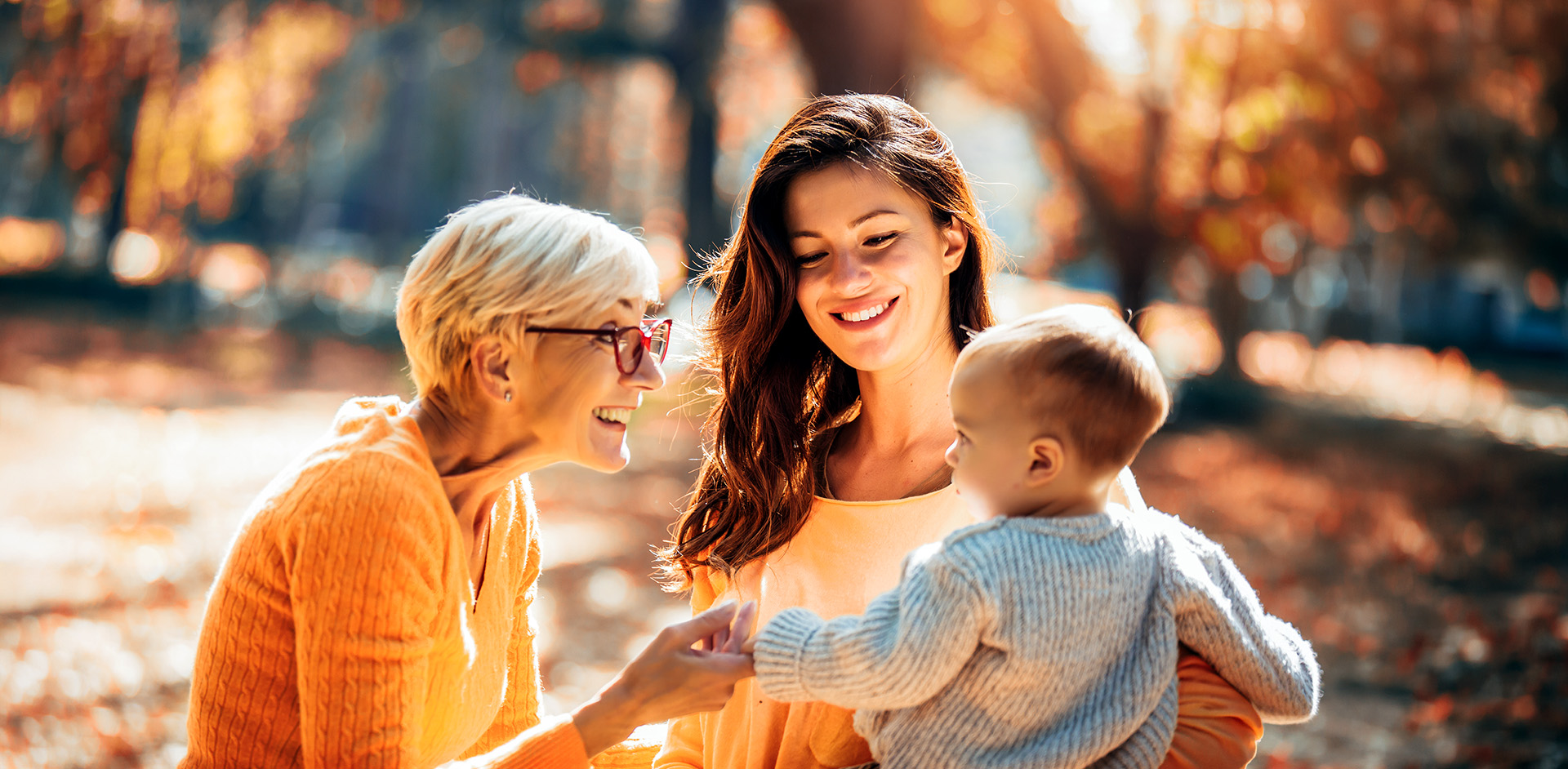 Grandmother and mother smiling at baby in autumn park.