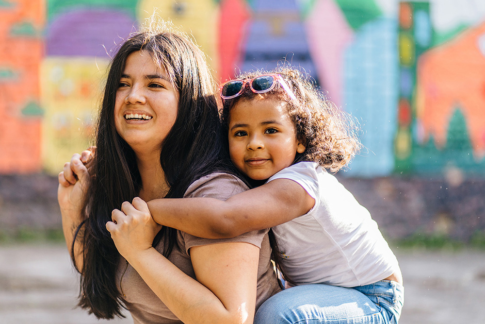 Enjoying ecuadorian mother playing and carrying young preschooler dark-skinned daughter on her shoulders while walking together in playground. Blurred children s drawing on background.